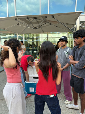 4 students are standing and speaking beneath an umbrella at the Emory Student Center as a Hatchery staff member reaches into a cooler.